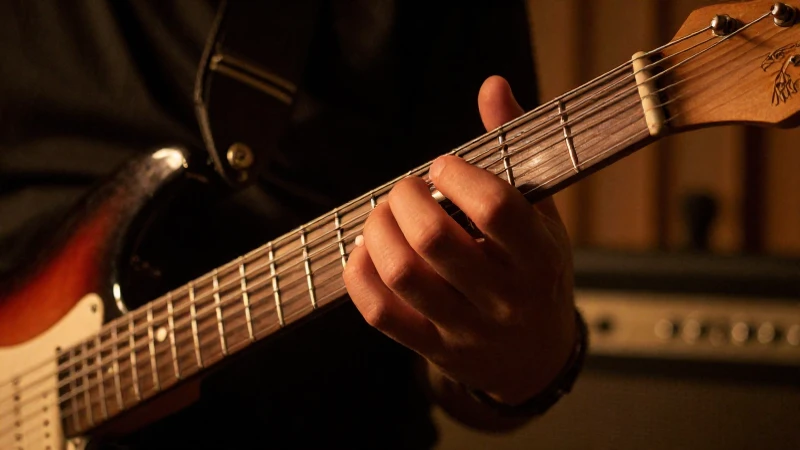 Primer plano de la mano de un guitarrista presionando las cuerdas de una guitarra eléctrica bajo luces cálidas.